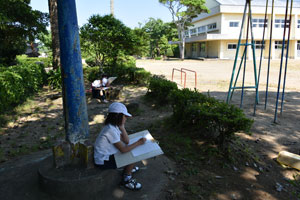 校庭で遊具施設や校舎の風景を描く児童